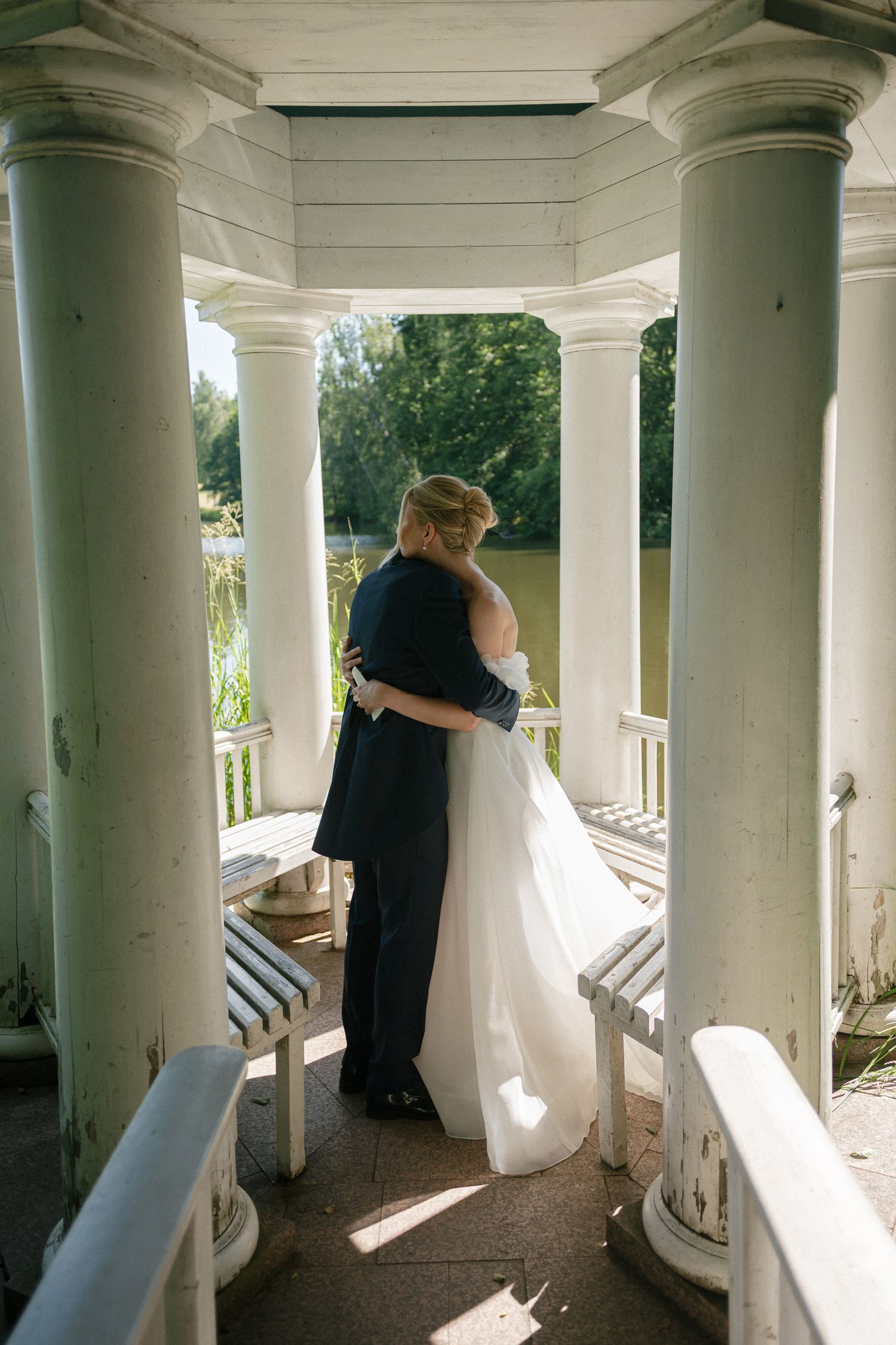 A bride and groom embrace in a white gazebo with columns, overlooking a serene garden pond on their wedding day.