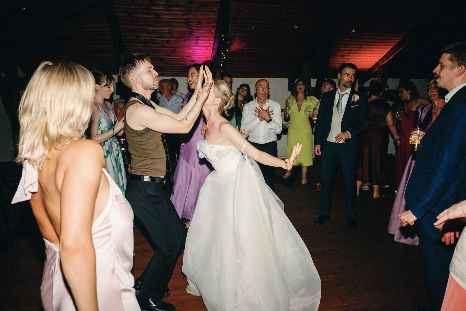 Bride in white wedding dress dancing joyfully with guests on the dance floor at a lively wedding reception.