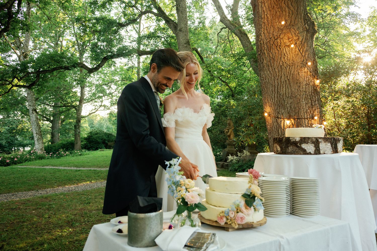Bride and groom cutting wedding cake outdoors under trees with string lights at elegant garden reception.
