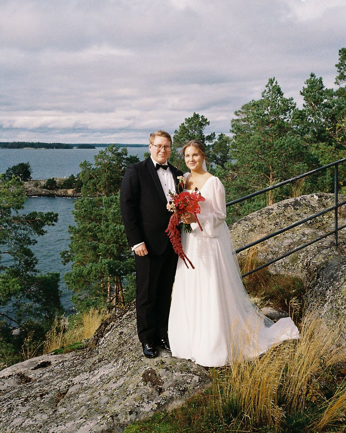 Bride and groom pose on rocky cliff overlooking scenic Scandinavian archipelago, she holds red bouquet, he wears black tuxedo.