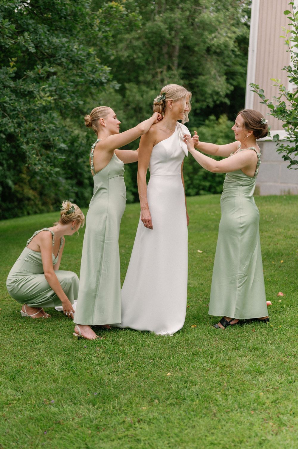 Bridesmaids in mint green dresses help style the bride's elegant white gown on a grassy lawn.
