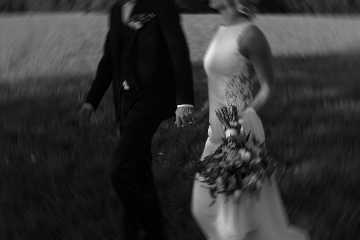 Artistic black and white capture of couple walking with wedding bouquet in field.