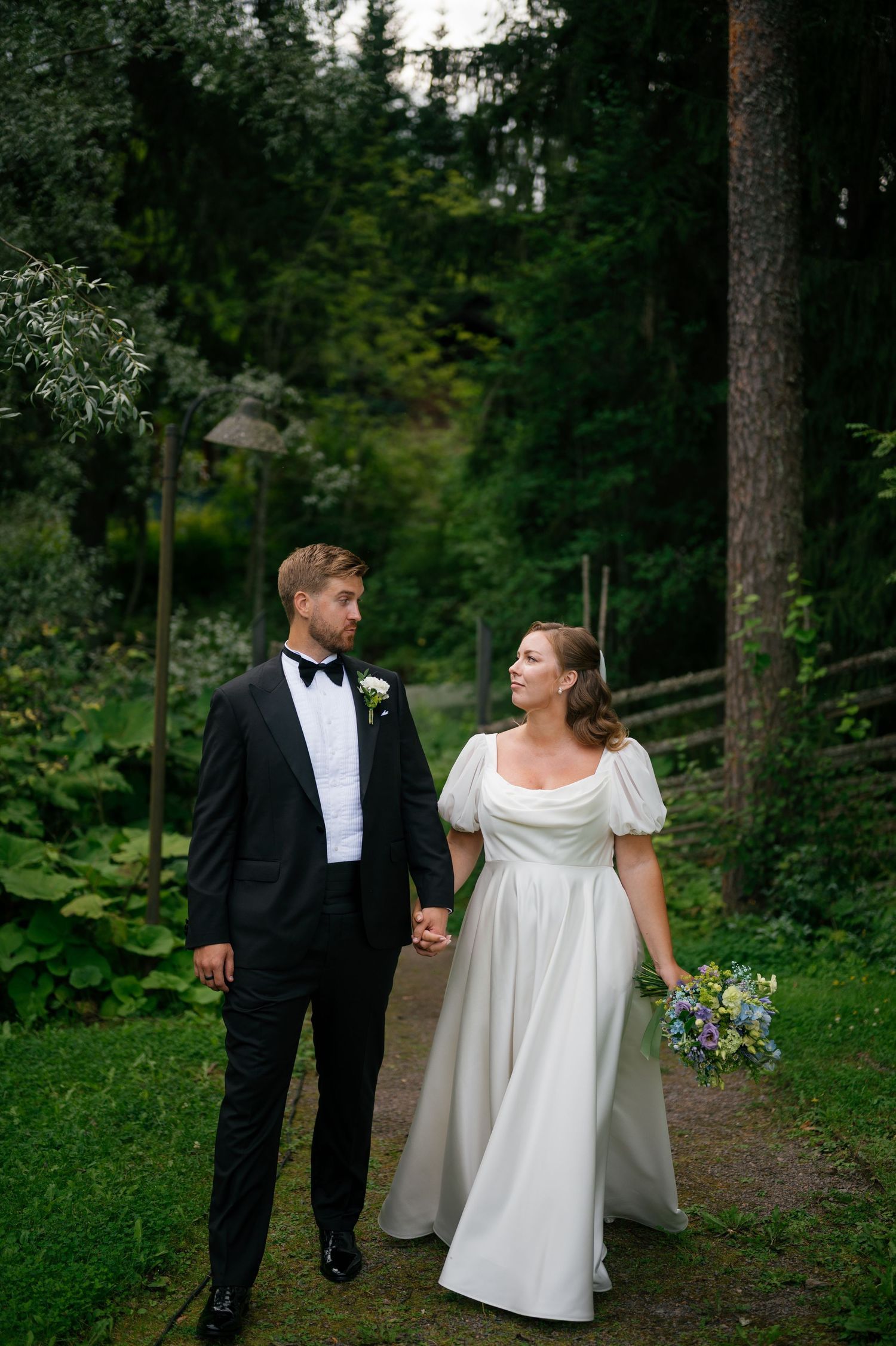 A couple walking hand in hand along a forest path in wedding attire.