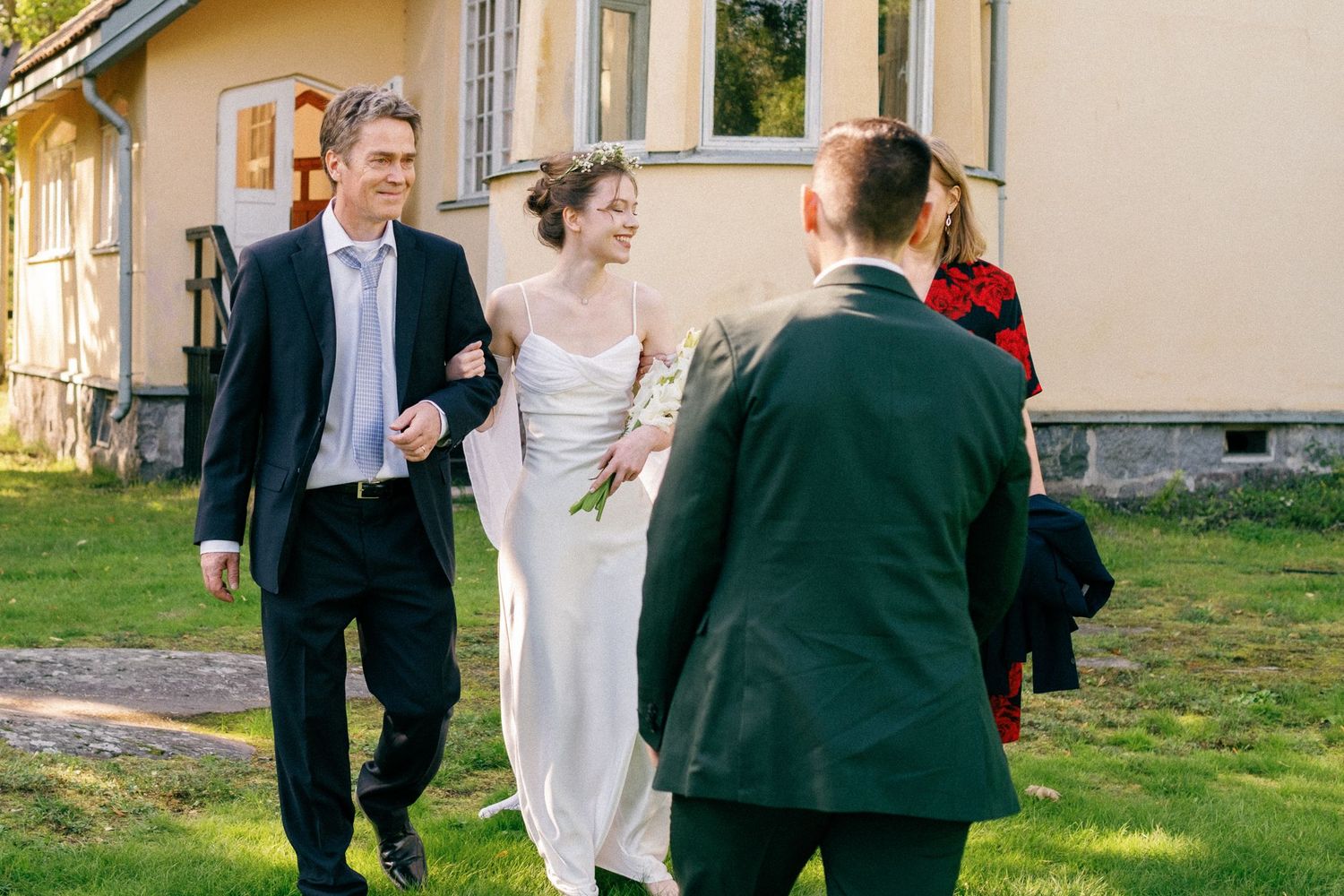 Wedding guests gather on a sunny lawn outside a stately yellow house for an outdoor celebration.