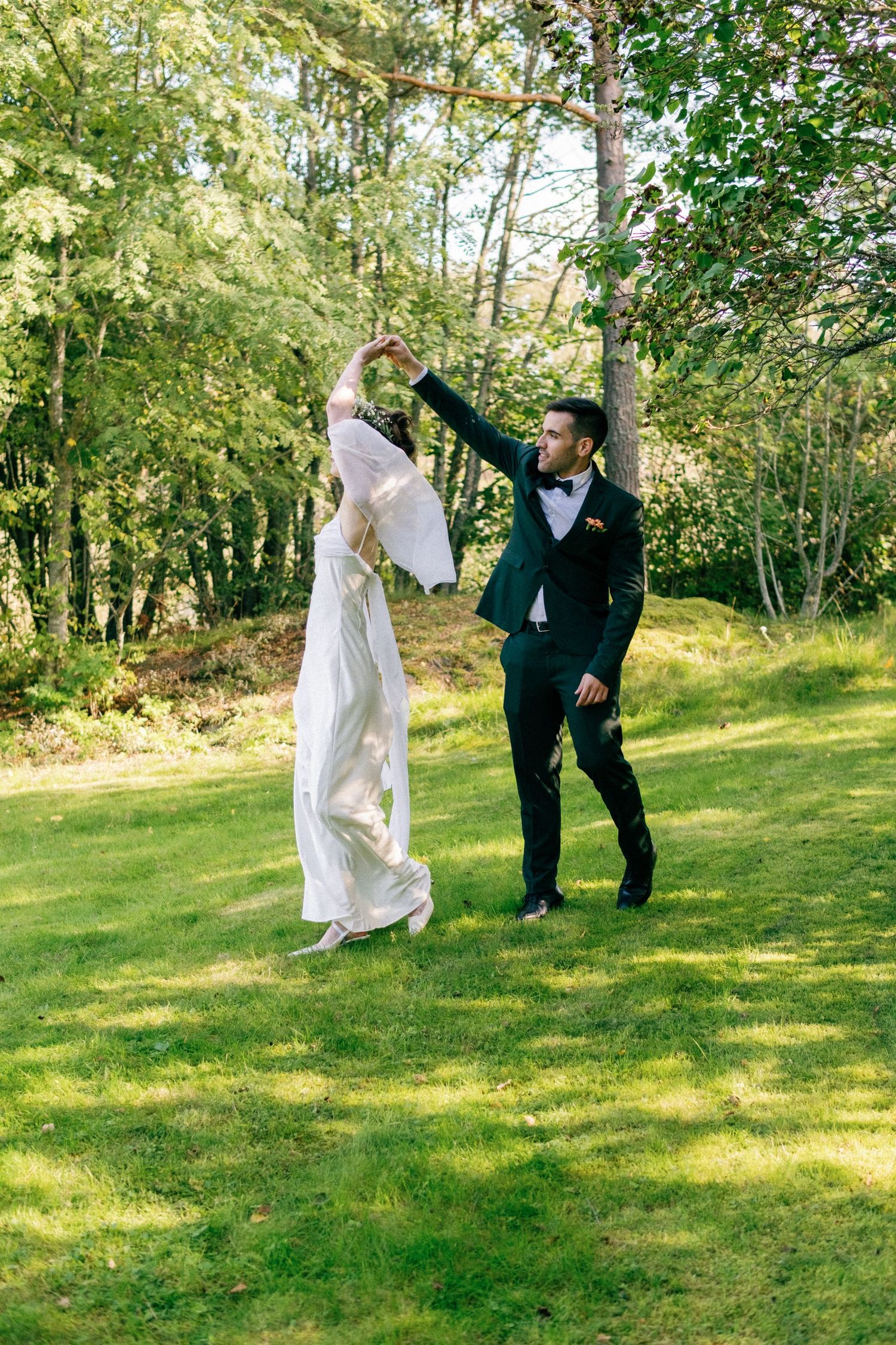 Wedding couple dances playfully on green grass with trees in the background.