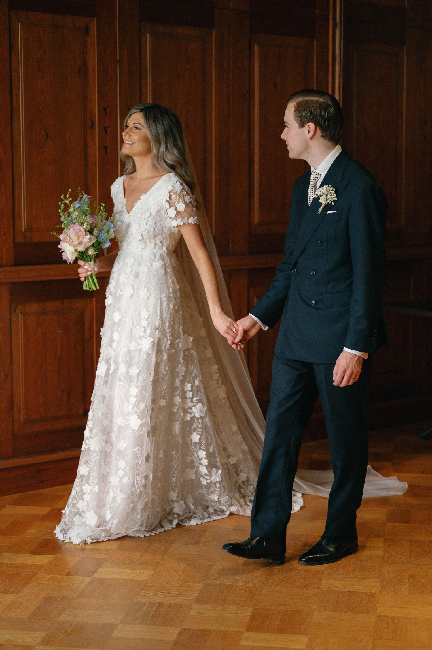 A couple in formal wedding attire dances together on a hardwood floor in front of wood-paneled walls.