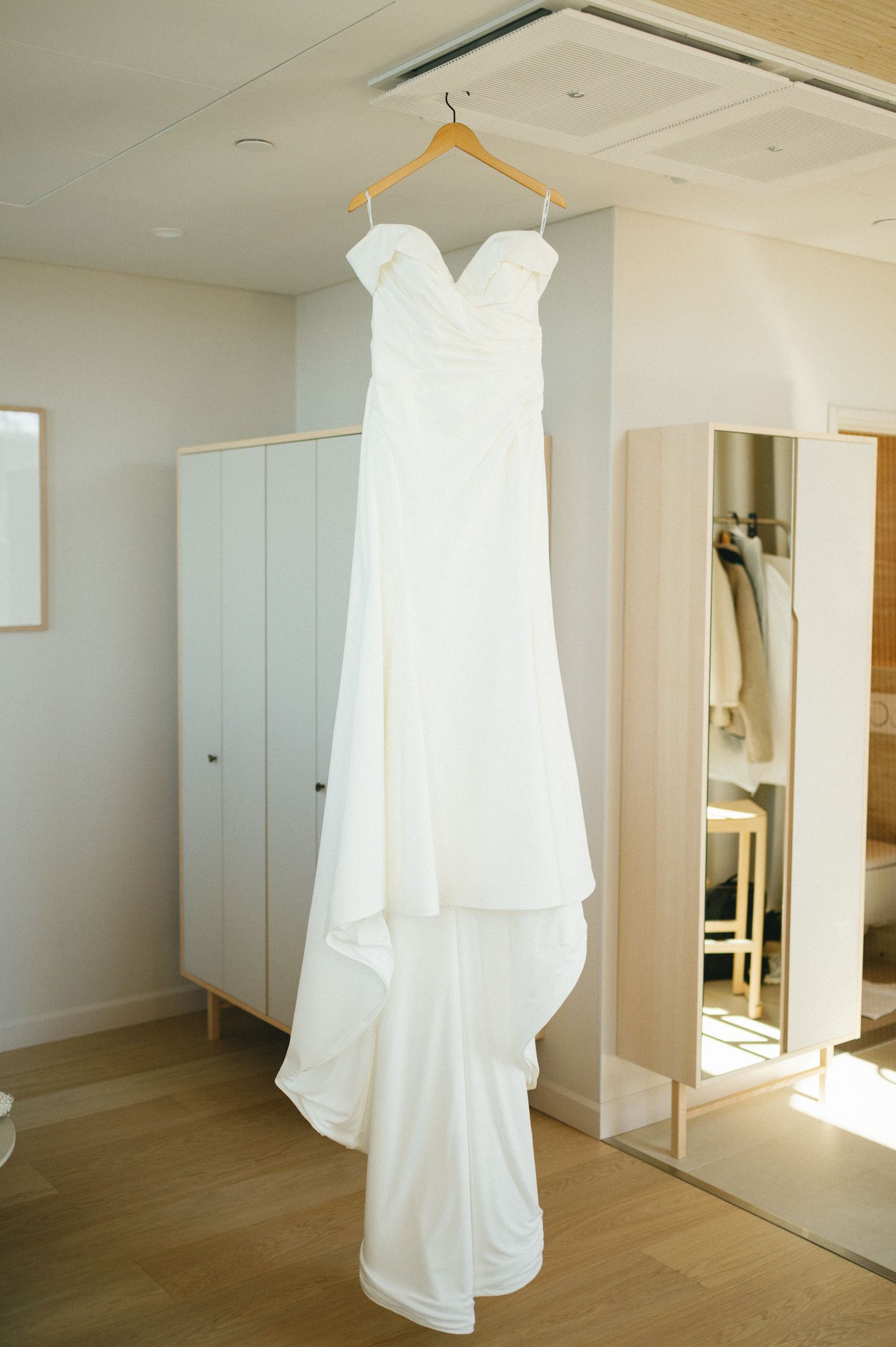 Elegant white strapless wedding dress with sweetheart neckline hanging on wooden hanger in bright modern room.