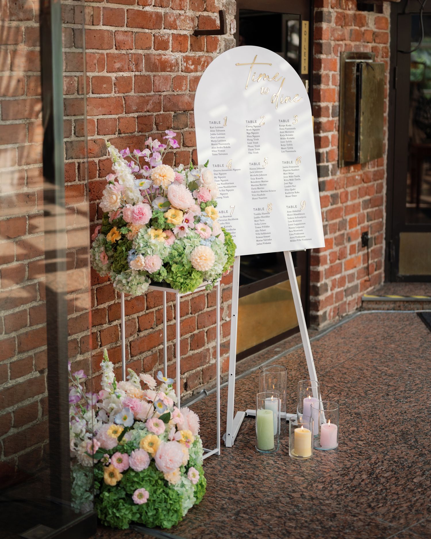 Elegant wedding seating chart on white arch easel with pastel floral arrangements and candles against brick wall.