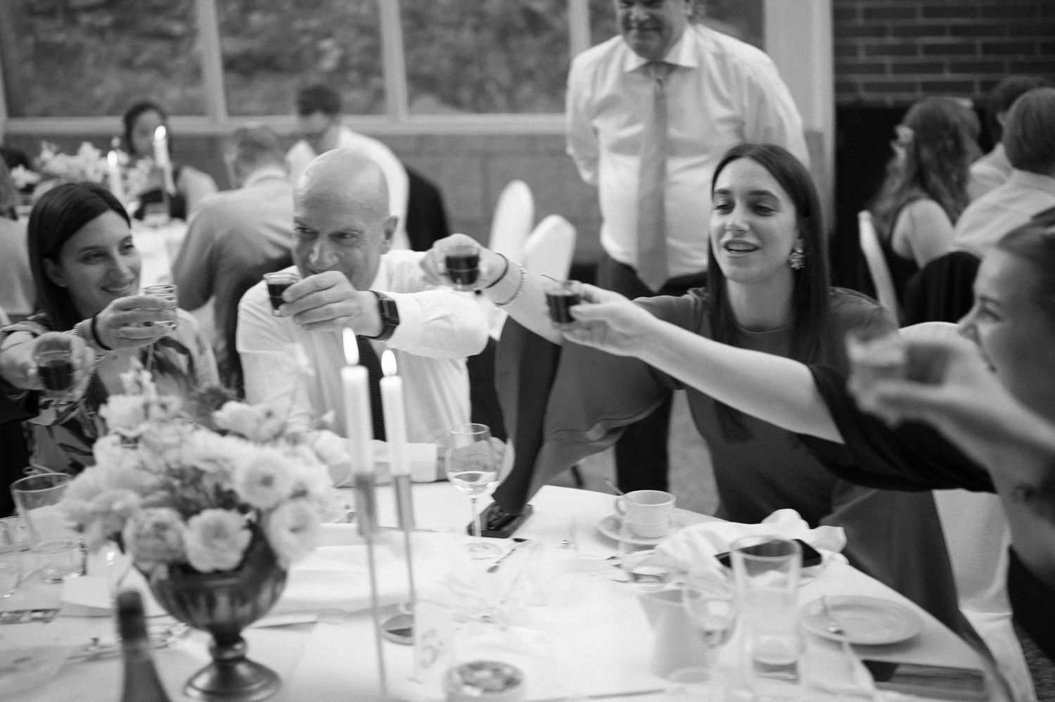 Black and white wedding reception toast with guests clinking glasses at elegantly decorated table with floral centerpiece.