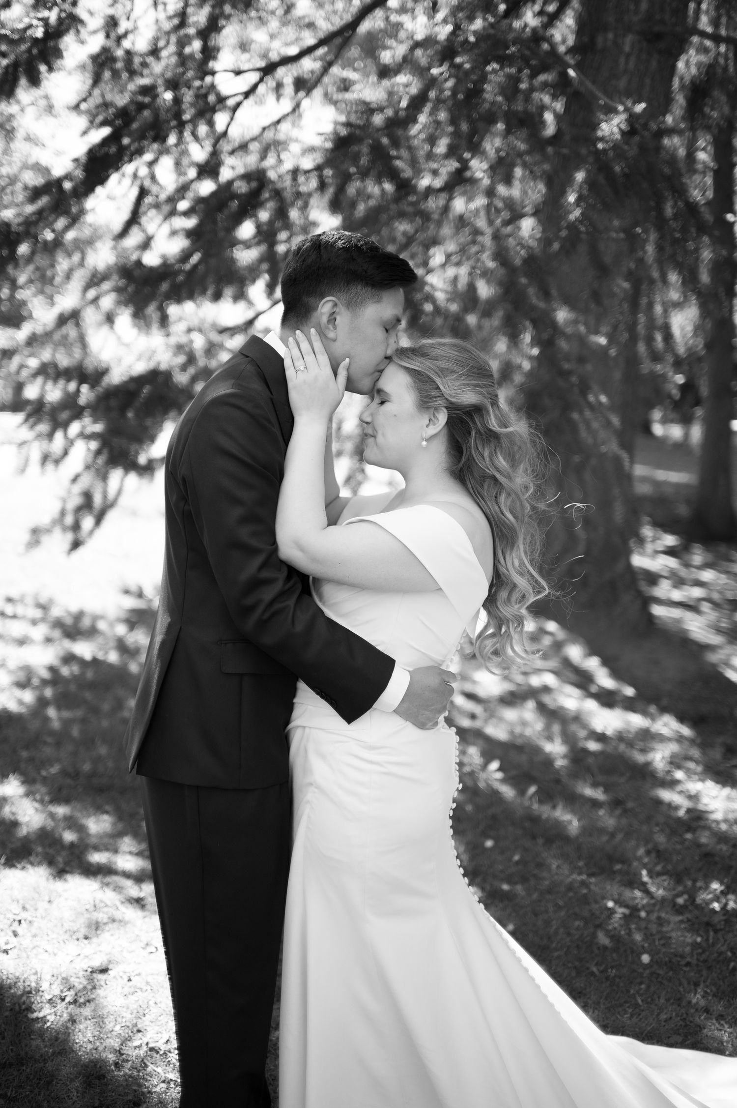 Black and white photo of a bride and groom embracing and sharing a romantic moment under trees on their wedding day.