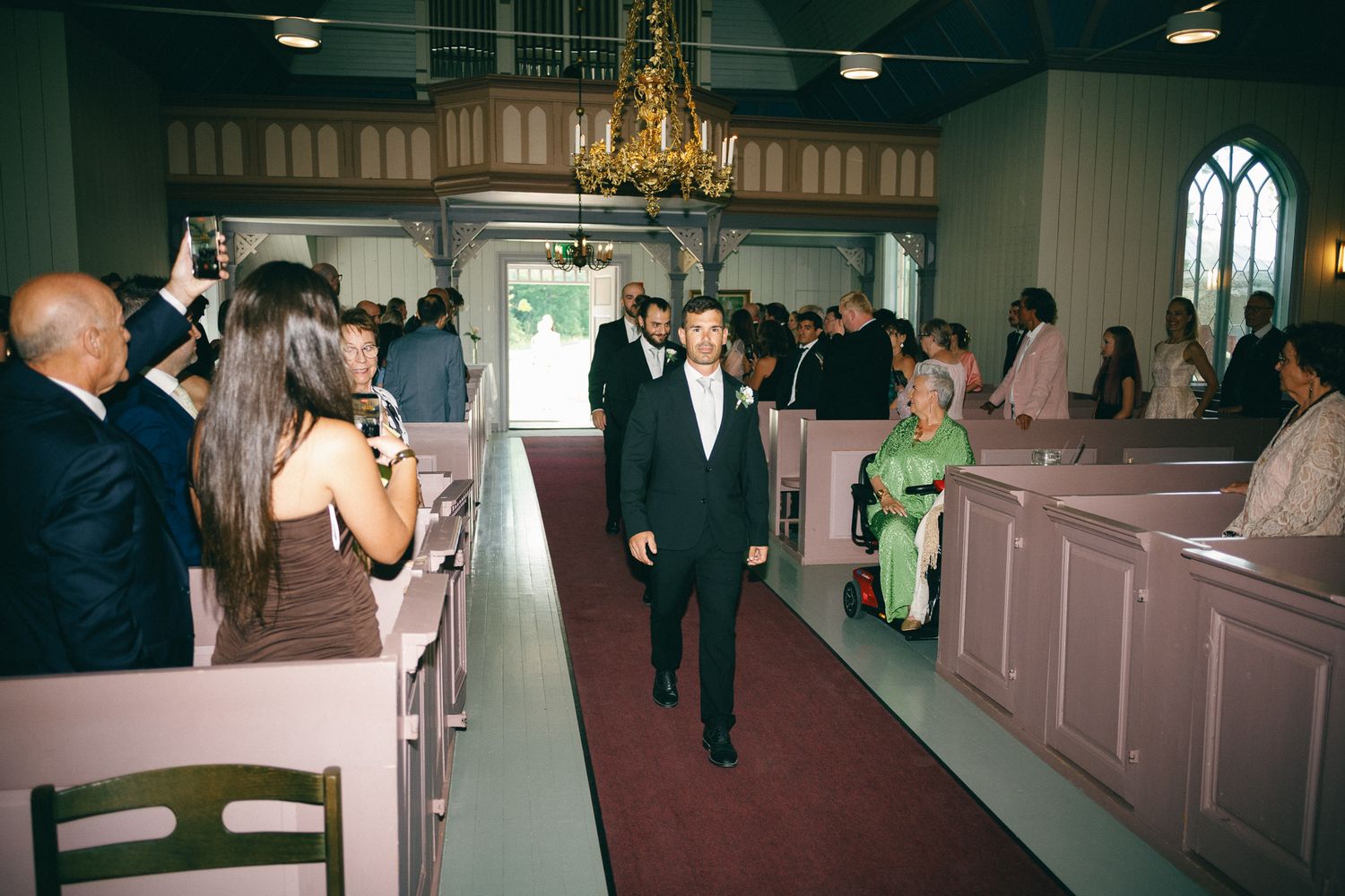 Groom walking down the aisle in a church wedding ceremony with guests seated in pink pews.