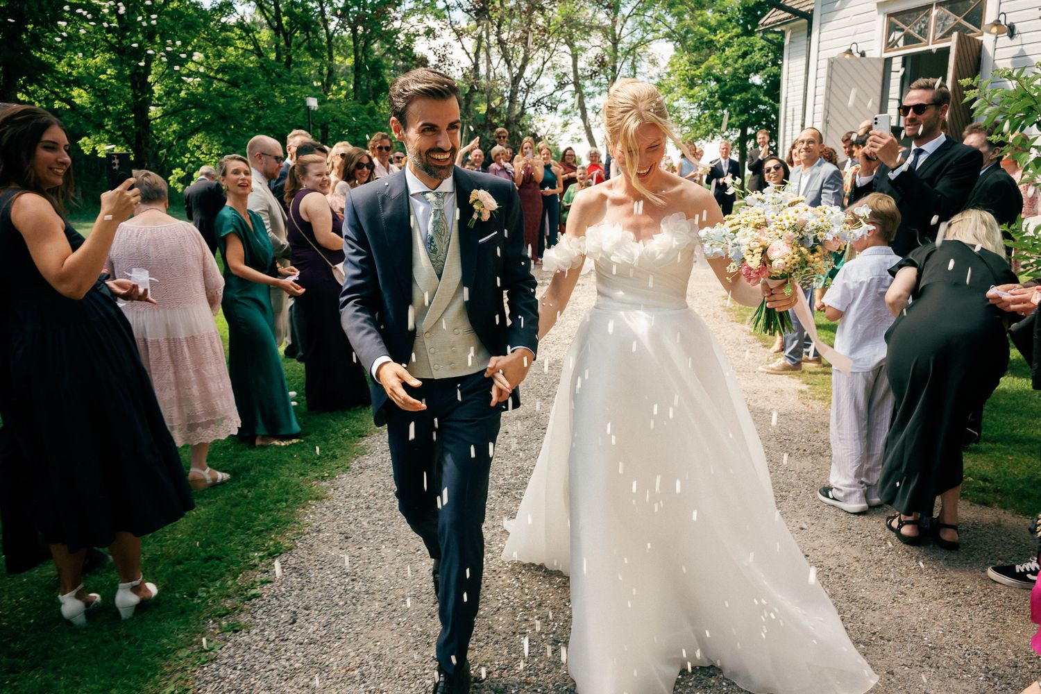 Bride and groom walk hand in hand through cheering guests throwing confetti at outdoor wedding ceremony.