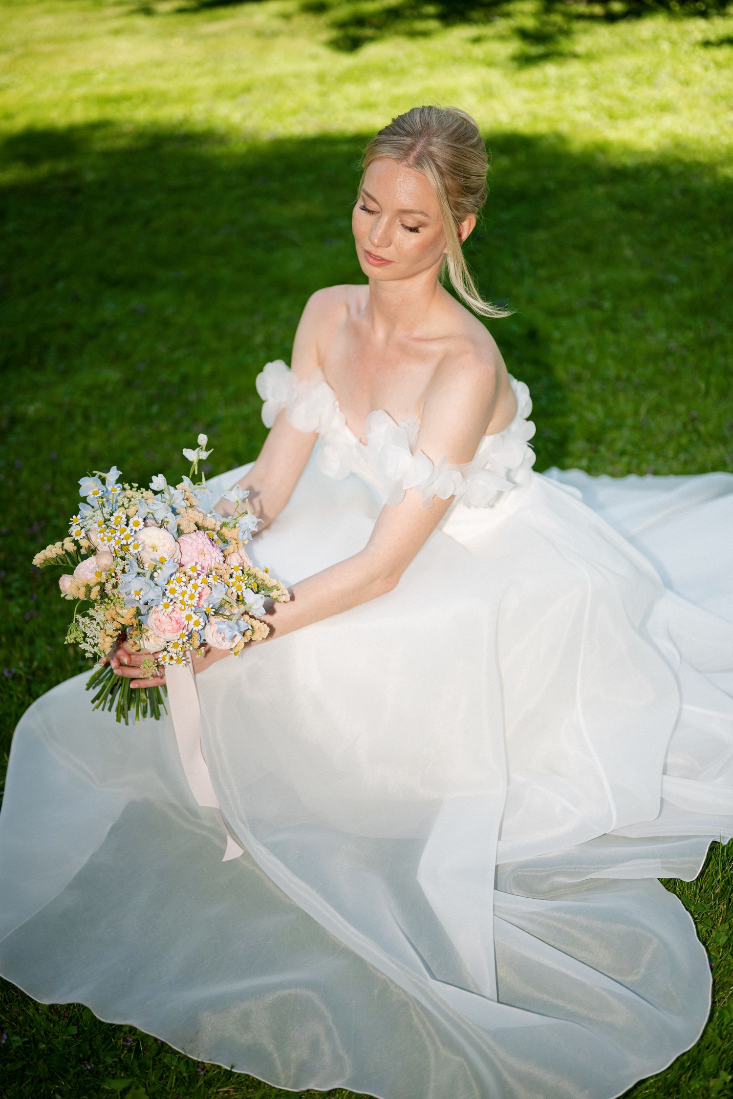 Bride in white off-shoulder gown sits on green grass holding a pastel floral bouquet outdoors.