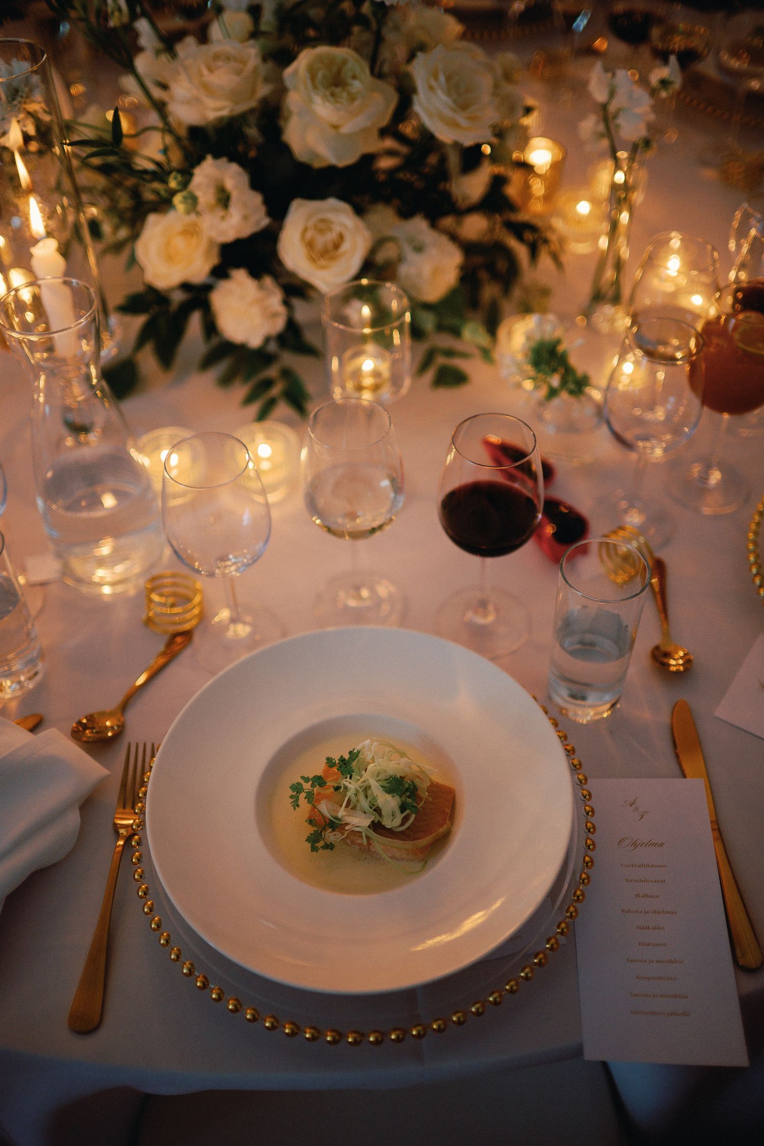 Elegant wedding reception place setting with gold cutlery, white soup bowl, red wine glass, and floral centerpiece with candles.
