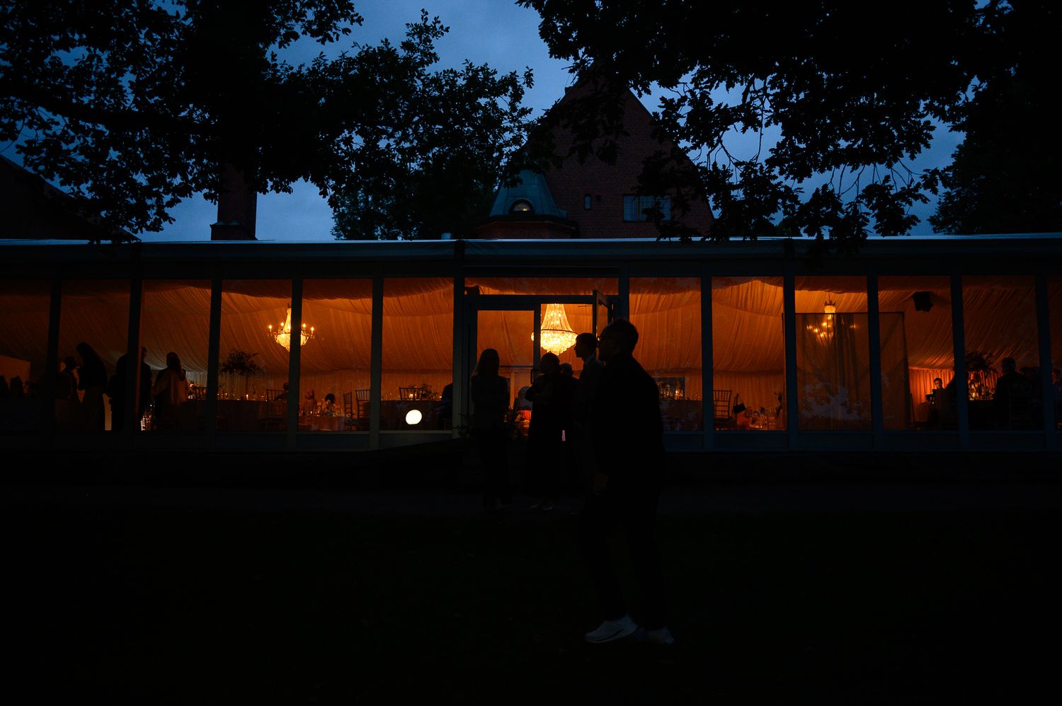 Illuminated glass pavilion glowing warmly at dusk with silhouetted guests gathered outside during an evening event.