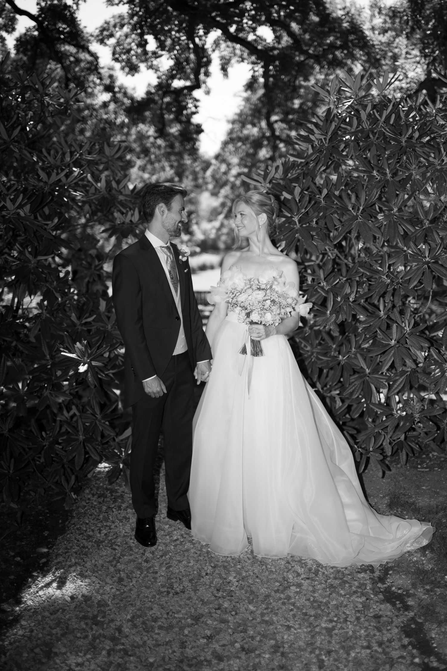 Black and white photo of a bride and groom walking hand in hand through a lush garden pathway on their wedding day.