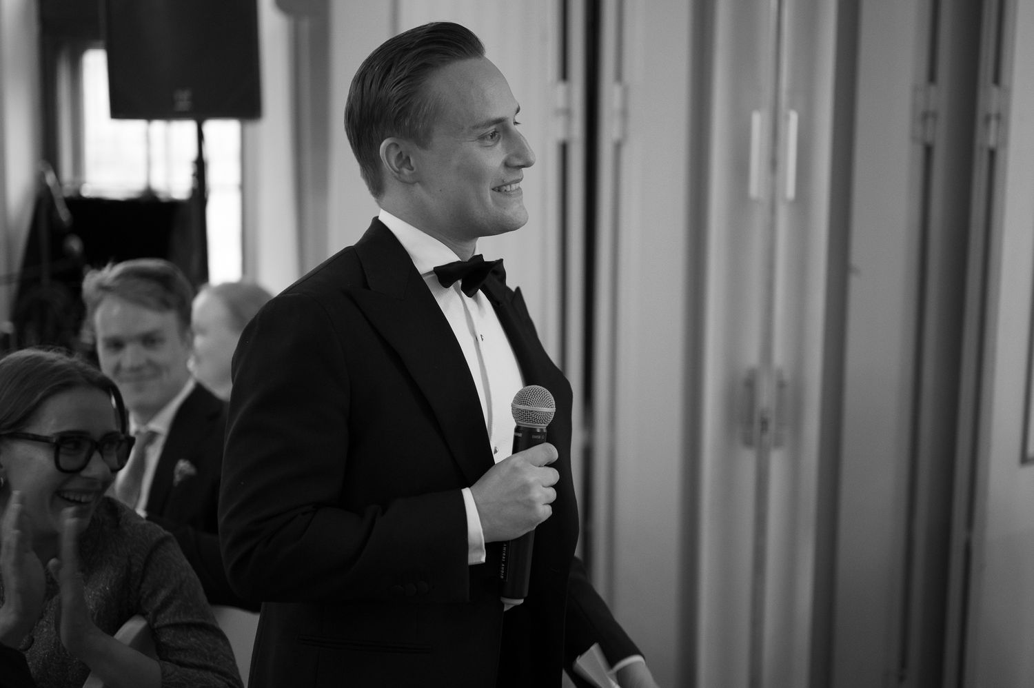 A person in a black tuxedo and bow tie smiles while speaking into a microphone at a formal event.
