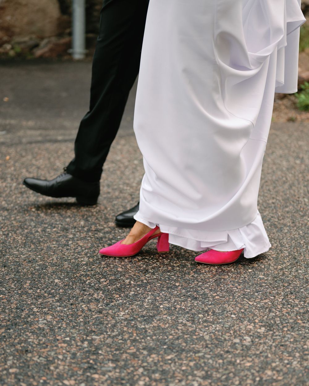 Red heels peek out beneath a flowing white wedding dress on a gravel path.