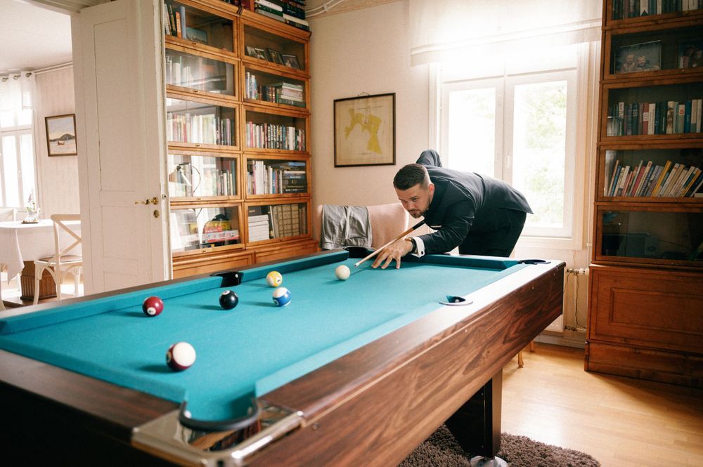Game room with blue pool table surrounded by wooden bookshelves and natural lighting.