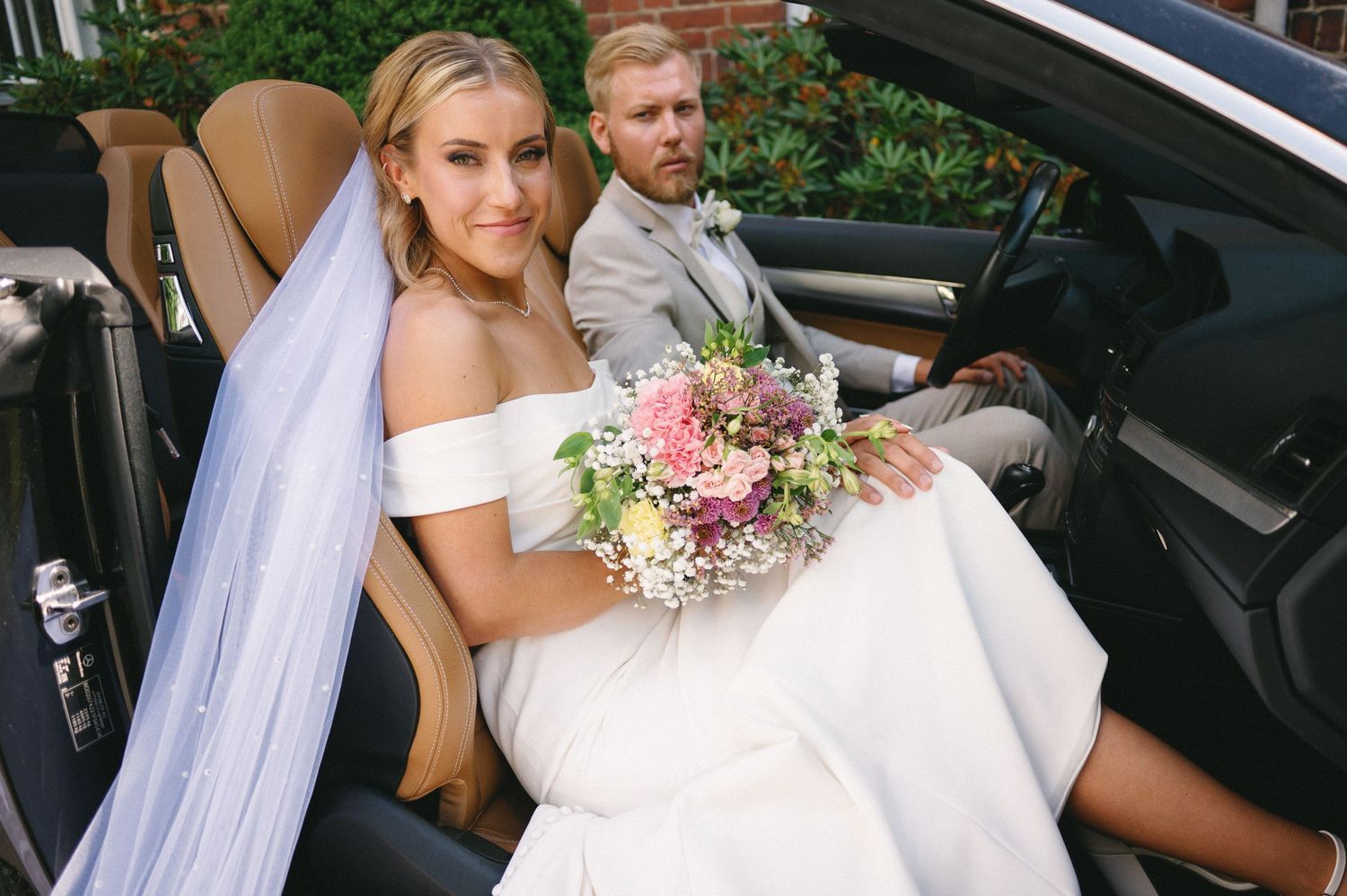 Bride with bouquet getting settled into car seat while wearing white gown.