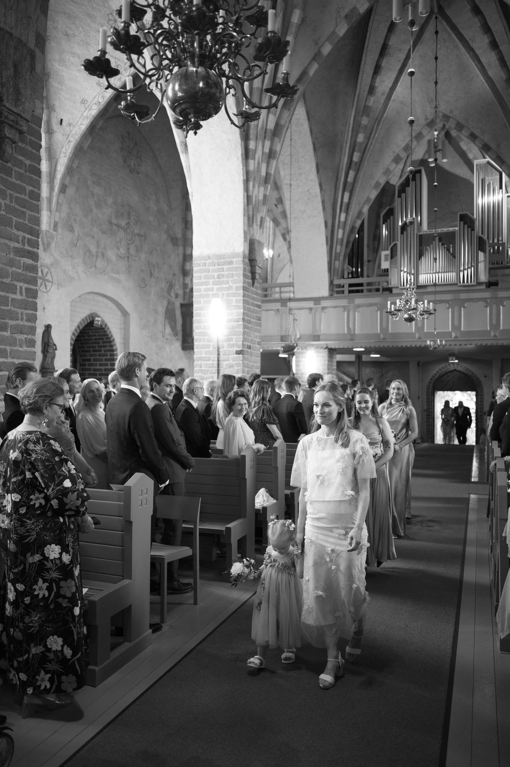 Wedding guests seated in wooden pews inside a historic brick church with vaulted ceilings and chandeliers.