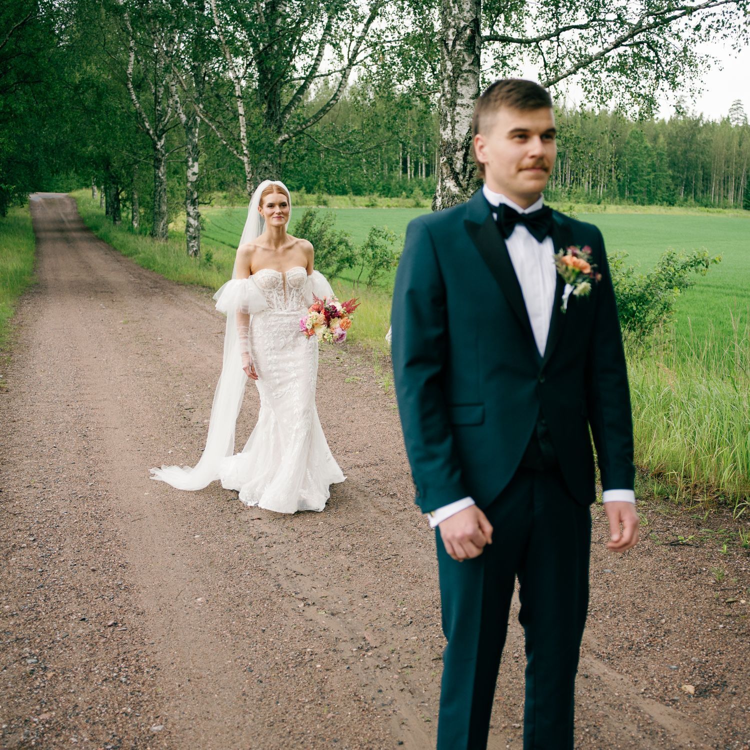 Wedding couple walking along a tree-lined dirt path through a green countryside.