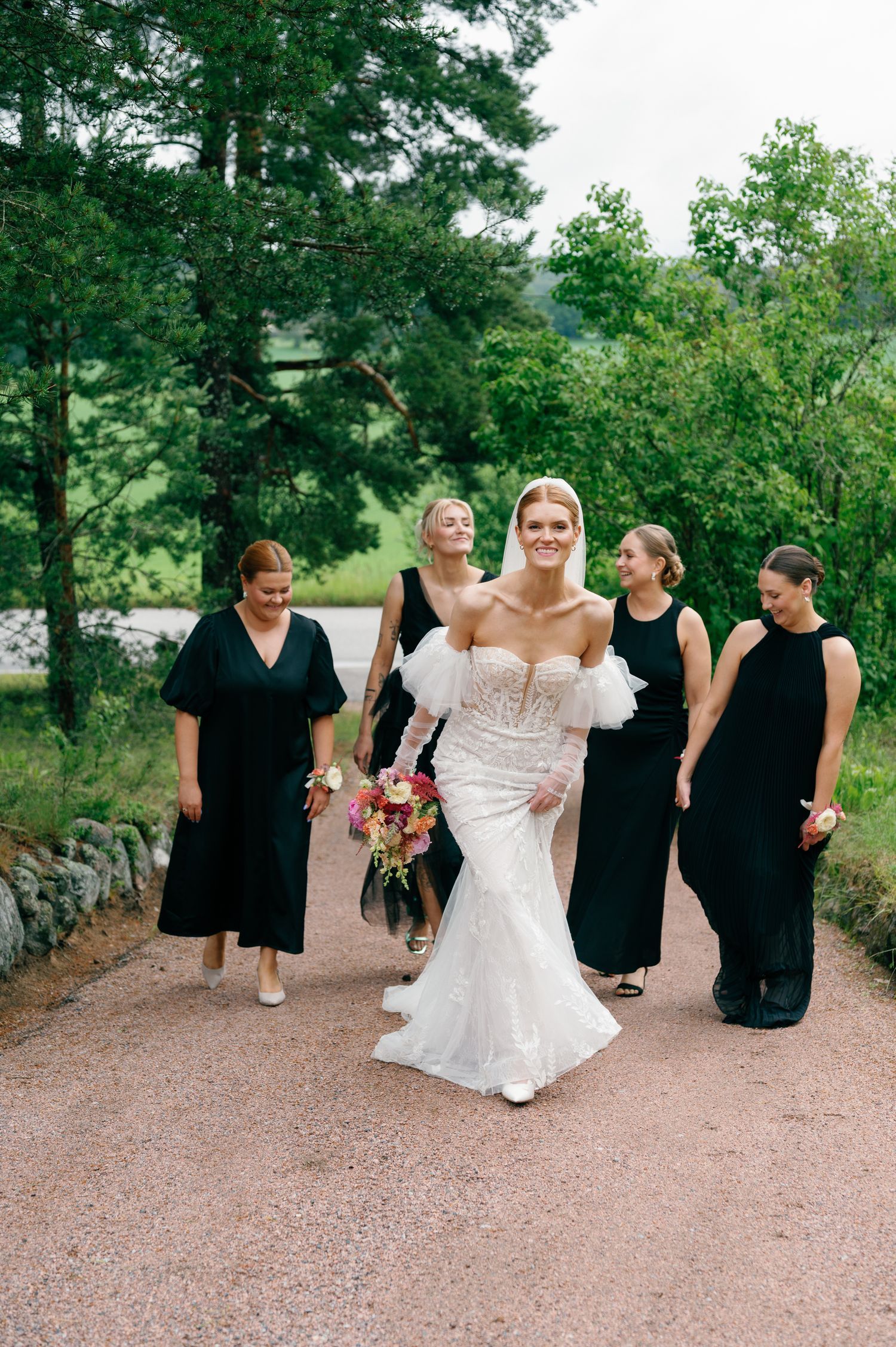 Bride in white gown walks with bridesmaids in black dresses along a garden path.