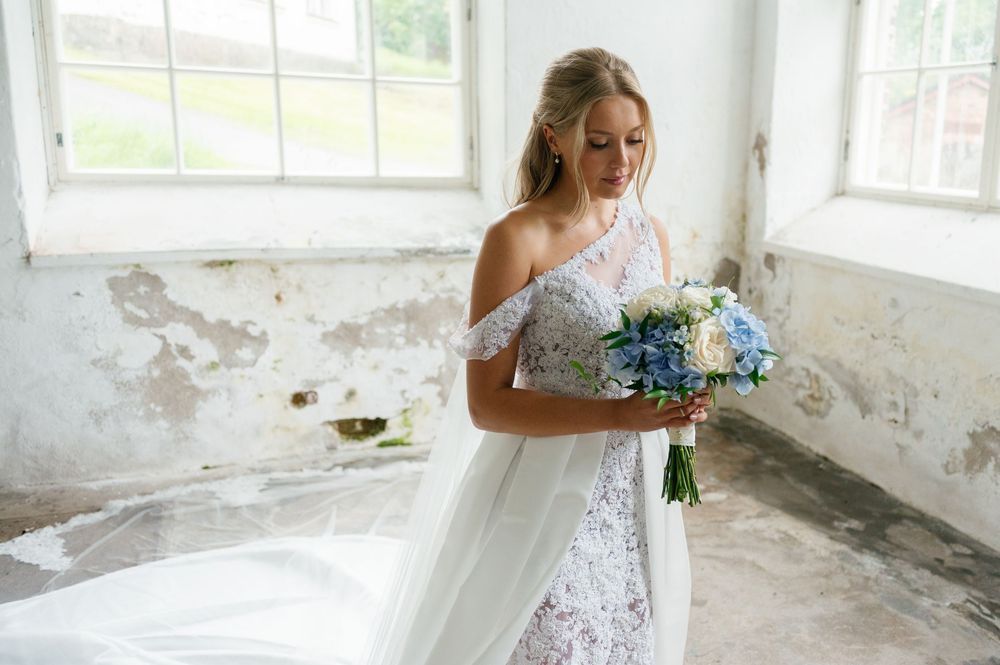 A bride holds a blue bouquet while posing in a rustic room with peeling walls and bright windows.