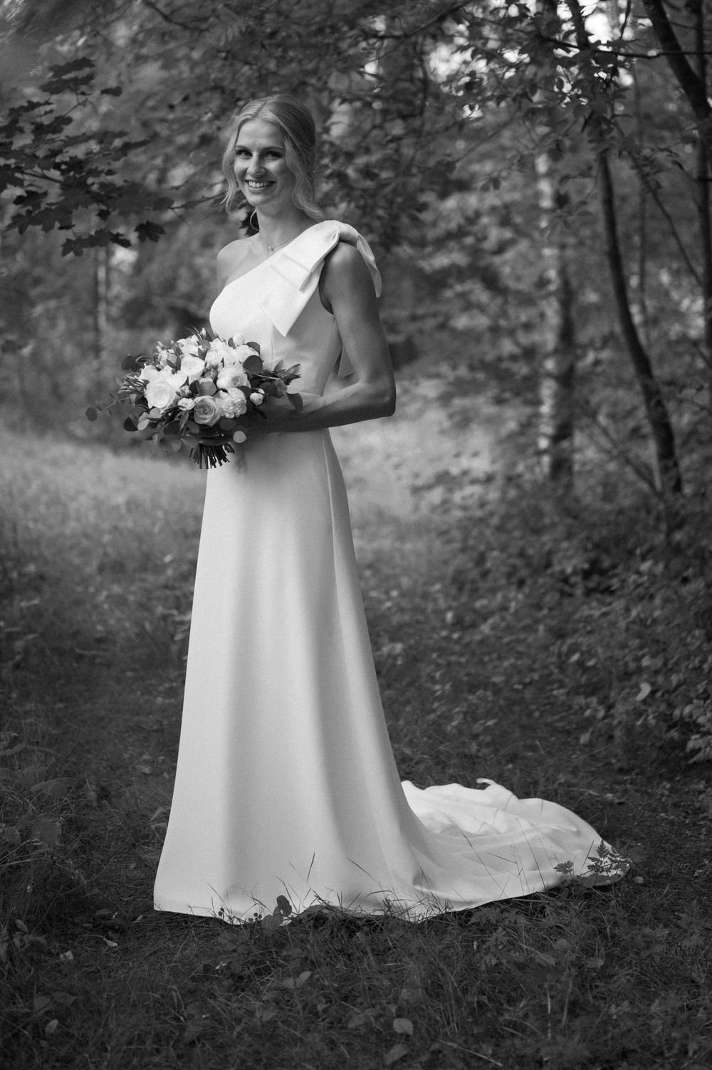 Bride in flowing white gown holding bouquet in wooded outdoor setting.