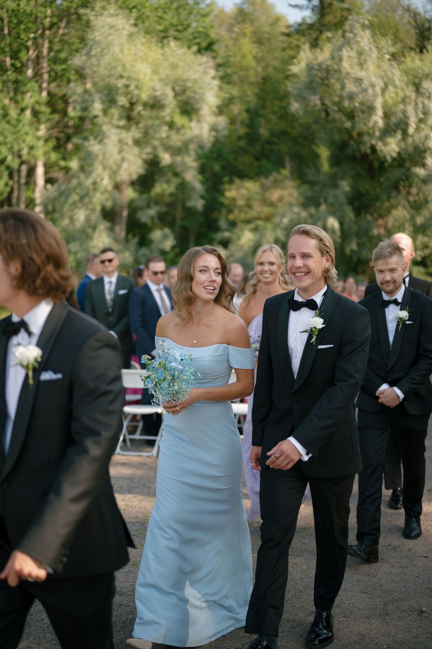 Wedding party members in formal attire walk together on a gravel path during the ceremony.