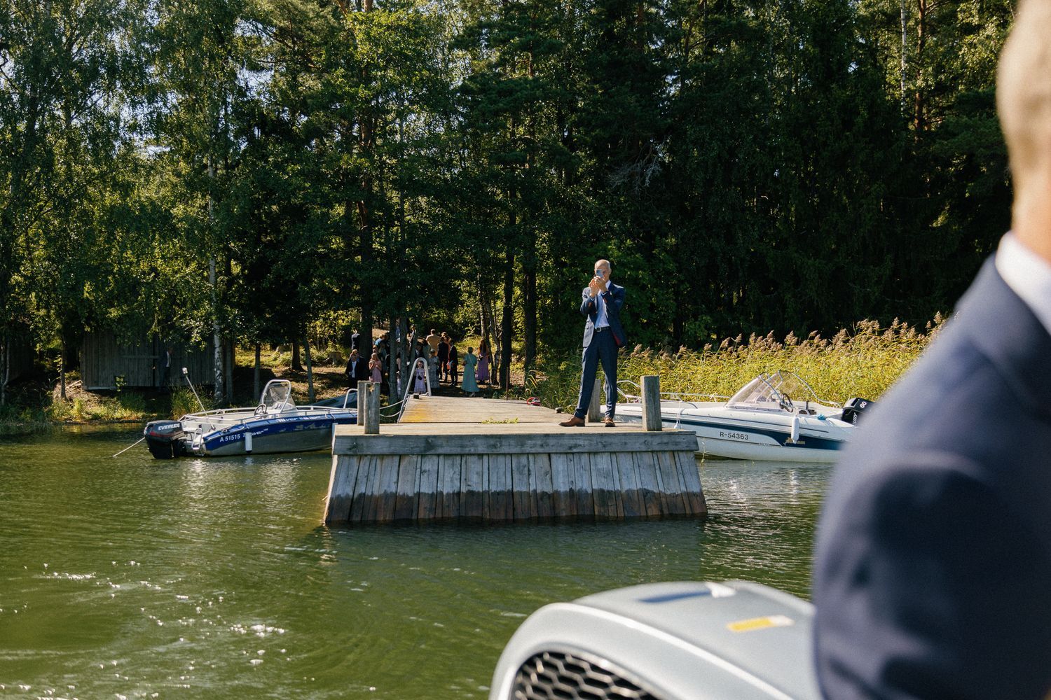 View of a wooden dock on a lake with boats and forest in the background.