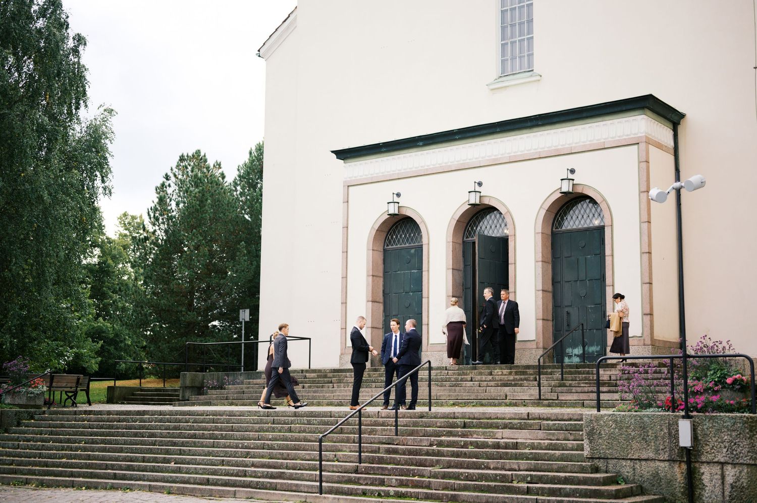 People walk near the entrance of a white church building with steps and arched doorways in a serene outdoor setting.