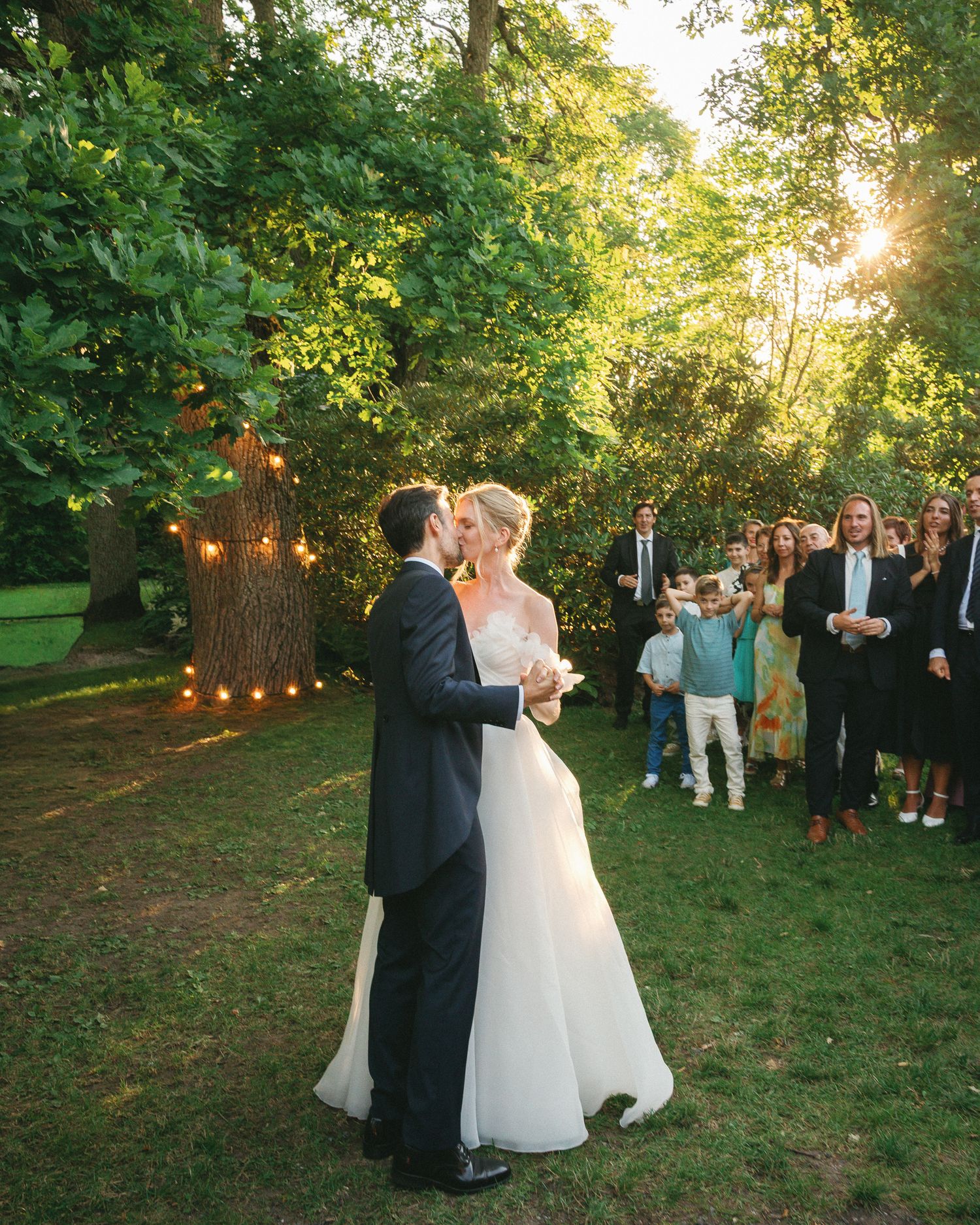 Bride and groom share first dance outdoors at golden hour wedding reception with guests watching among trees.