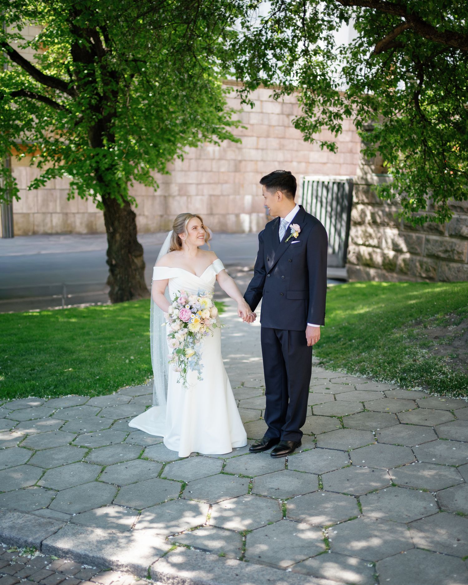 A bride and groom hold hands on a stone path surrounded by lush green trees on their wedding day.