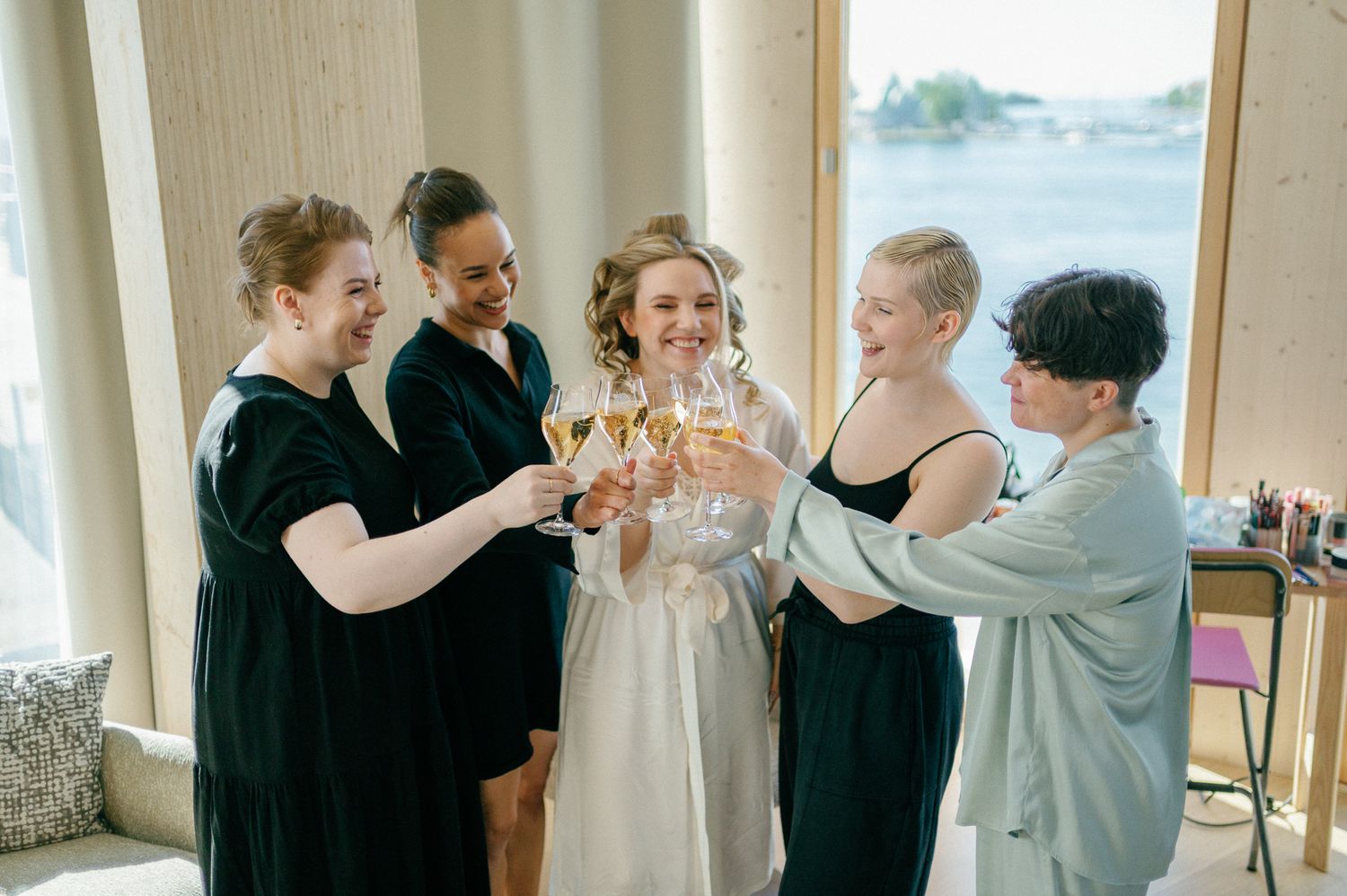 Bride in white robe toasting champagne with four bridesmaids in dark dresses near a waterfront window.
