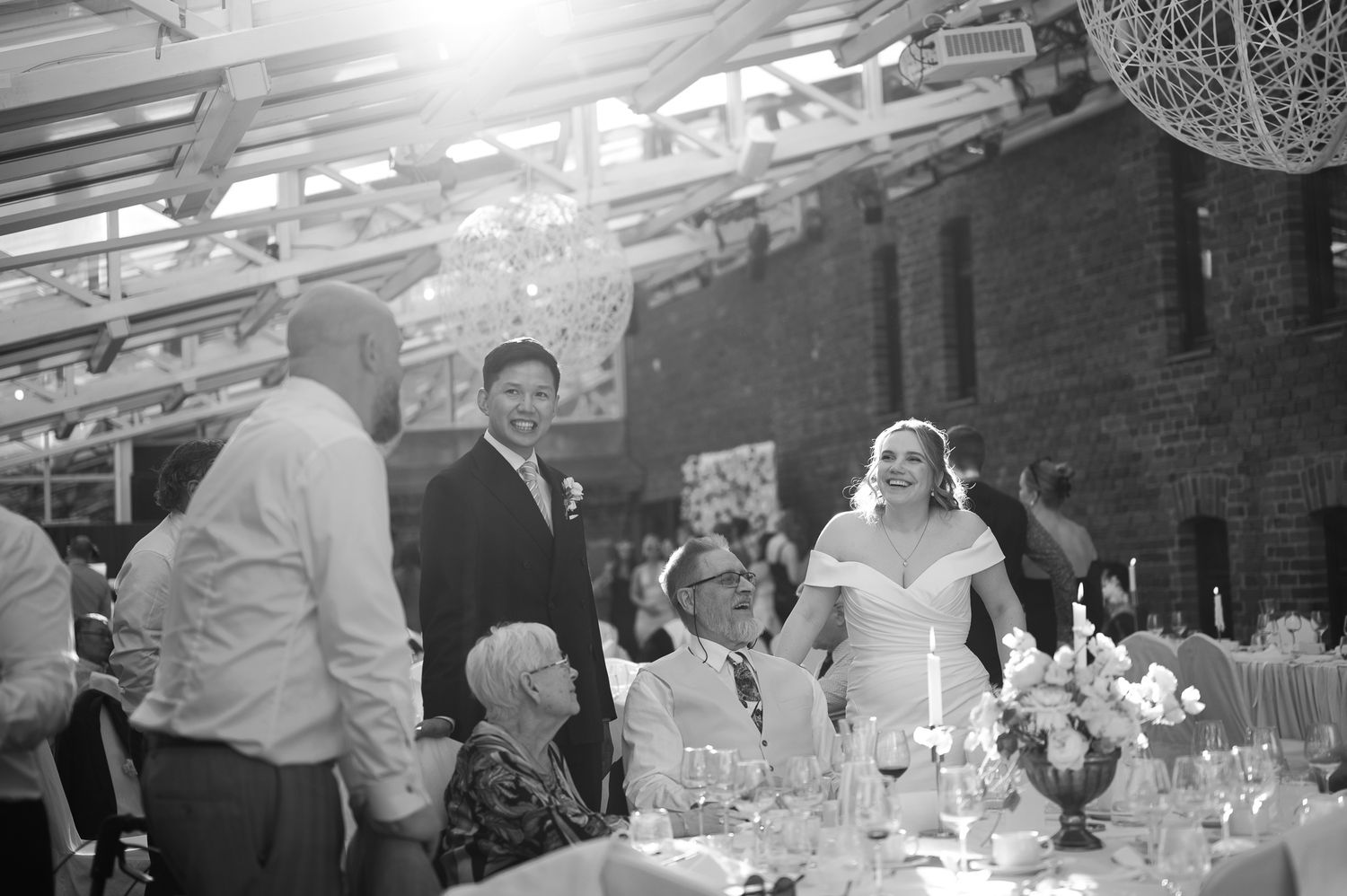 Black and white wedding reception photo showing bride and groom laughing with guests at elegantly decorated table.