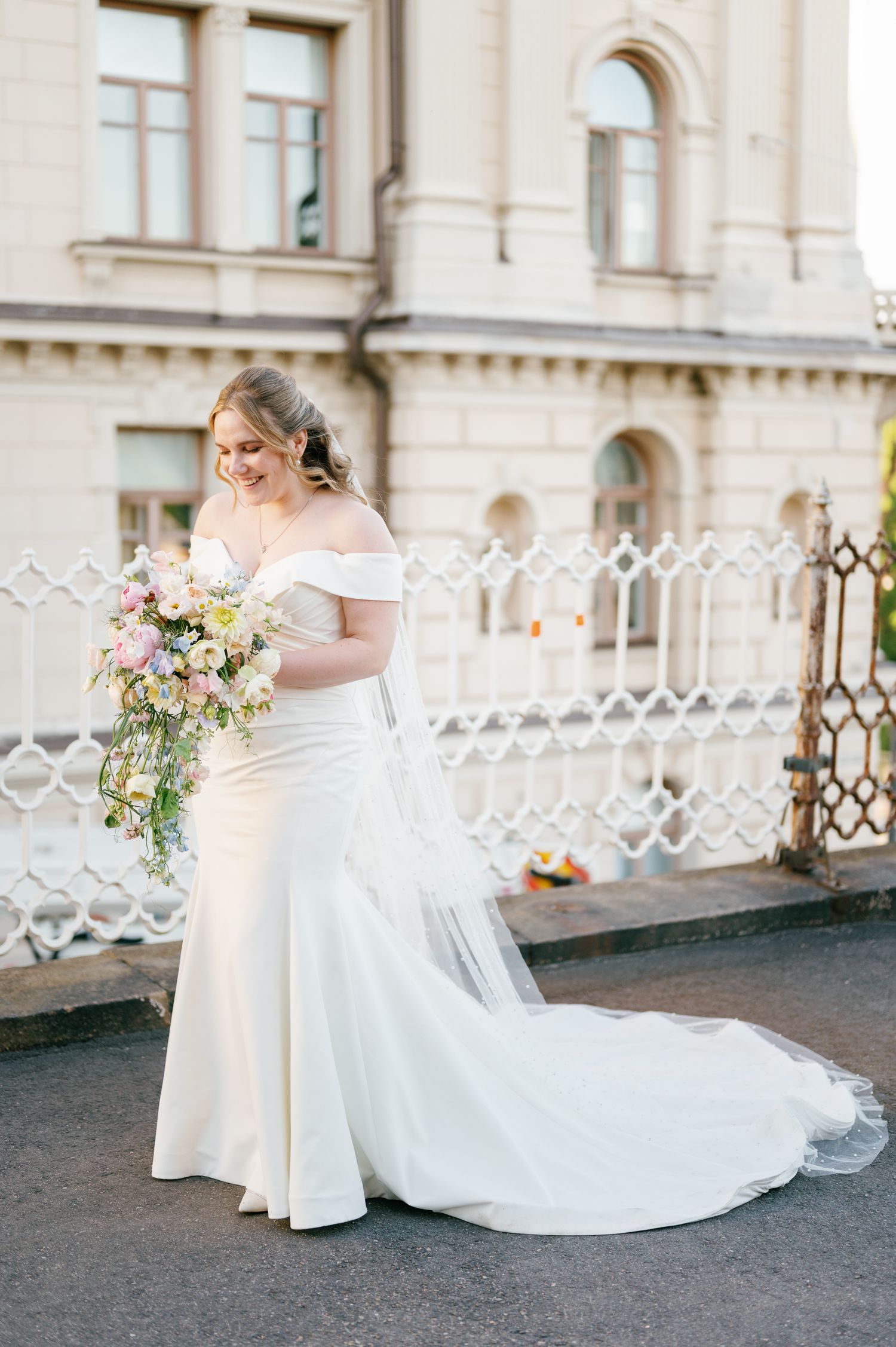 Bride in off-shoulder white mermaid gown holds pastel bouquet, standing before ornate building with iron fence.