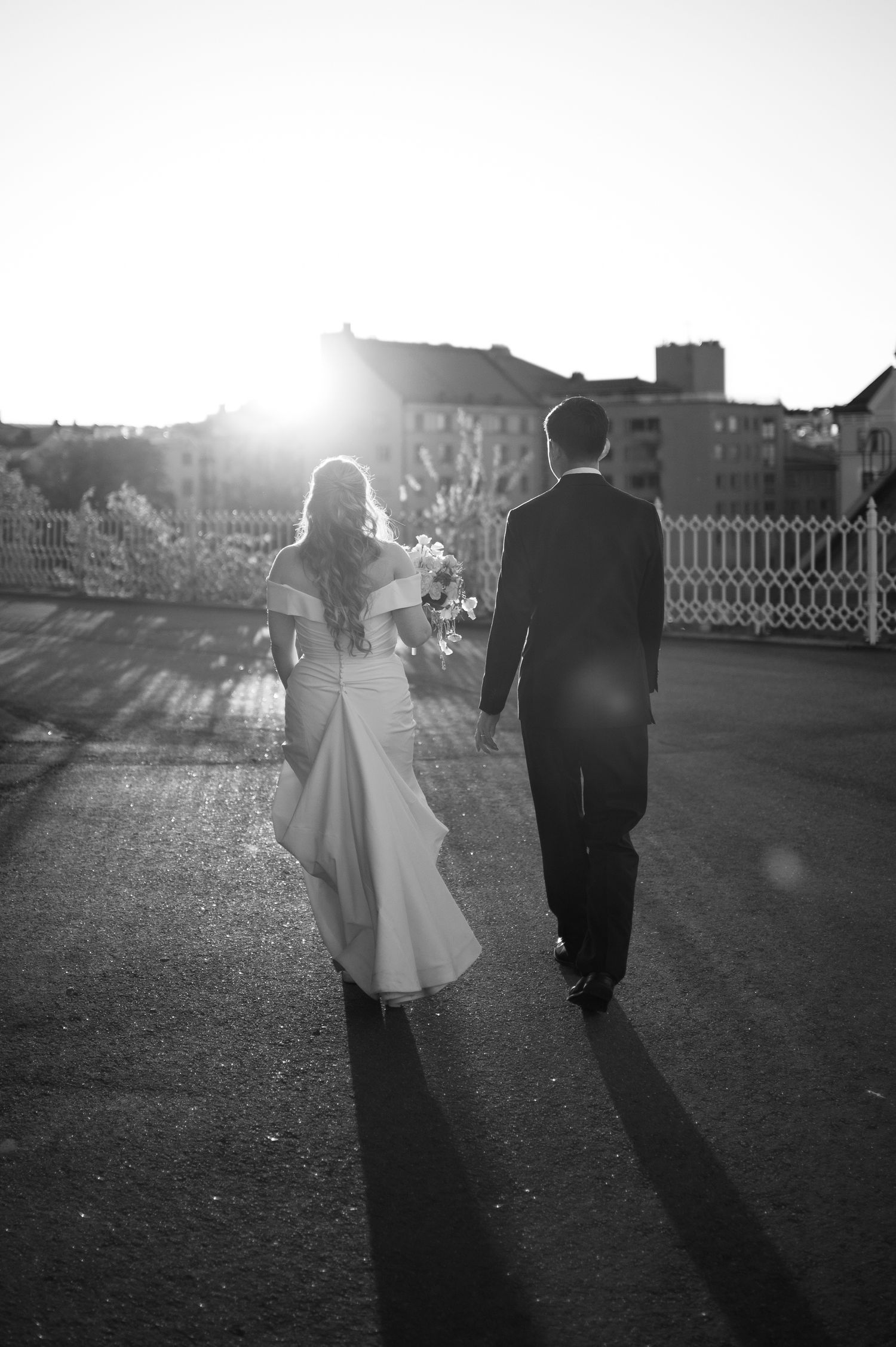 Black and white photo of a bride and groom walking hand in hand toward a sunlit horizon on their wedding day.