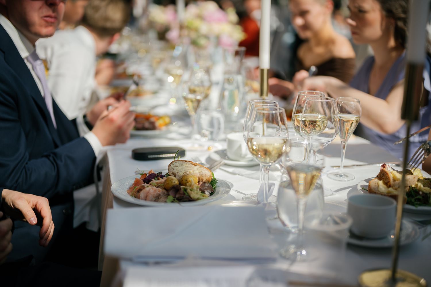 Elegant outdoor dinner party with guests seated at a long table adorned with wine glasses and gourmet dishes.
