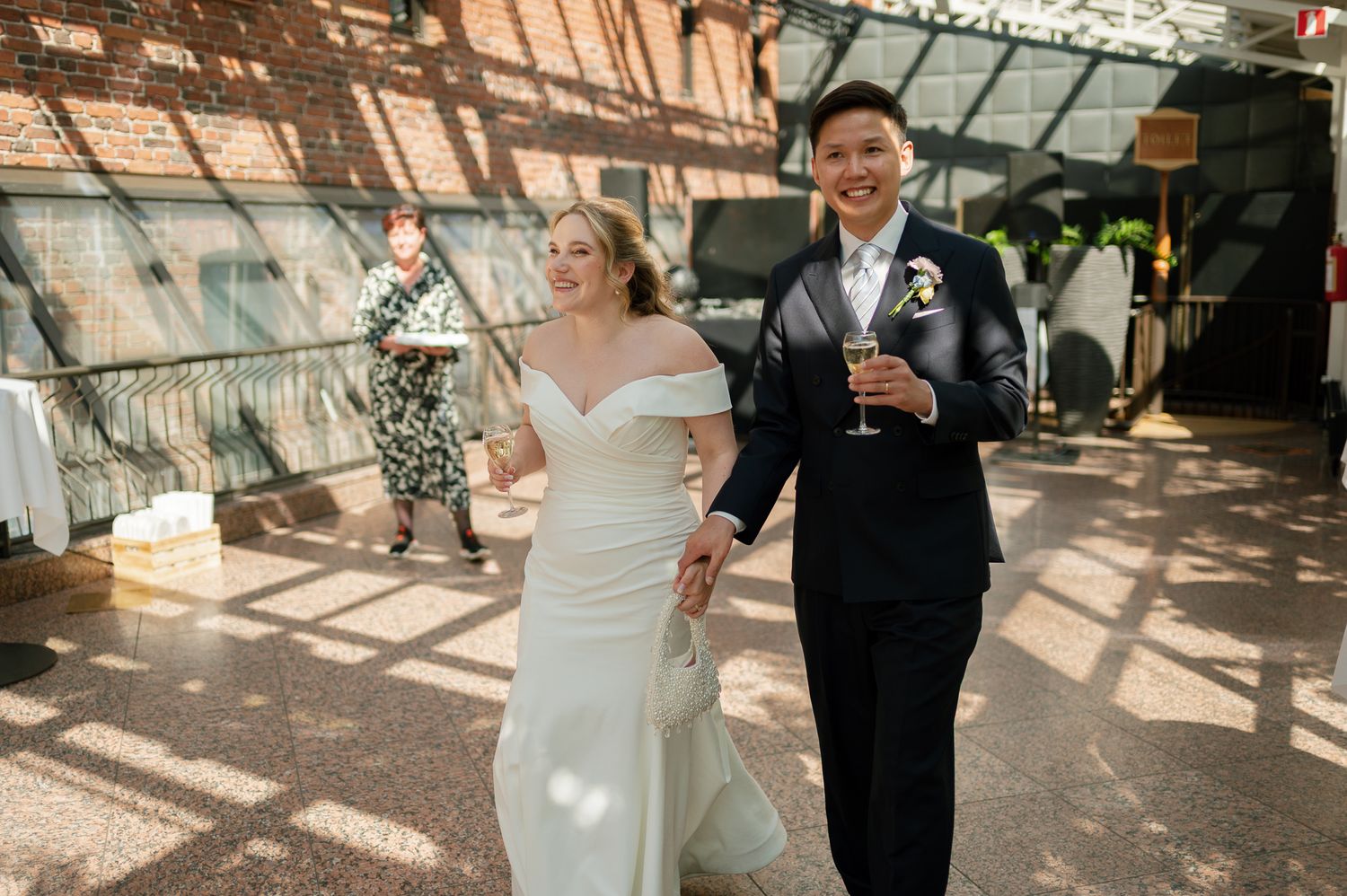 A bride in a white off-shoulder gown and groom in a black suit walk hand-in-hand at their elegant wedding reception.
