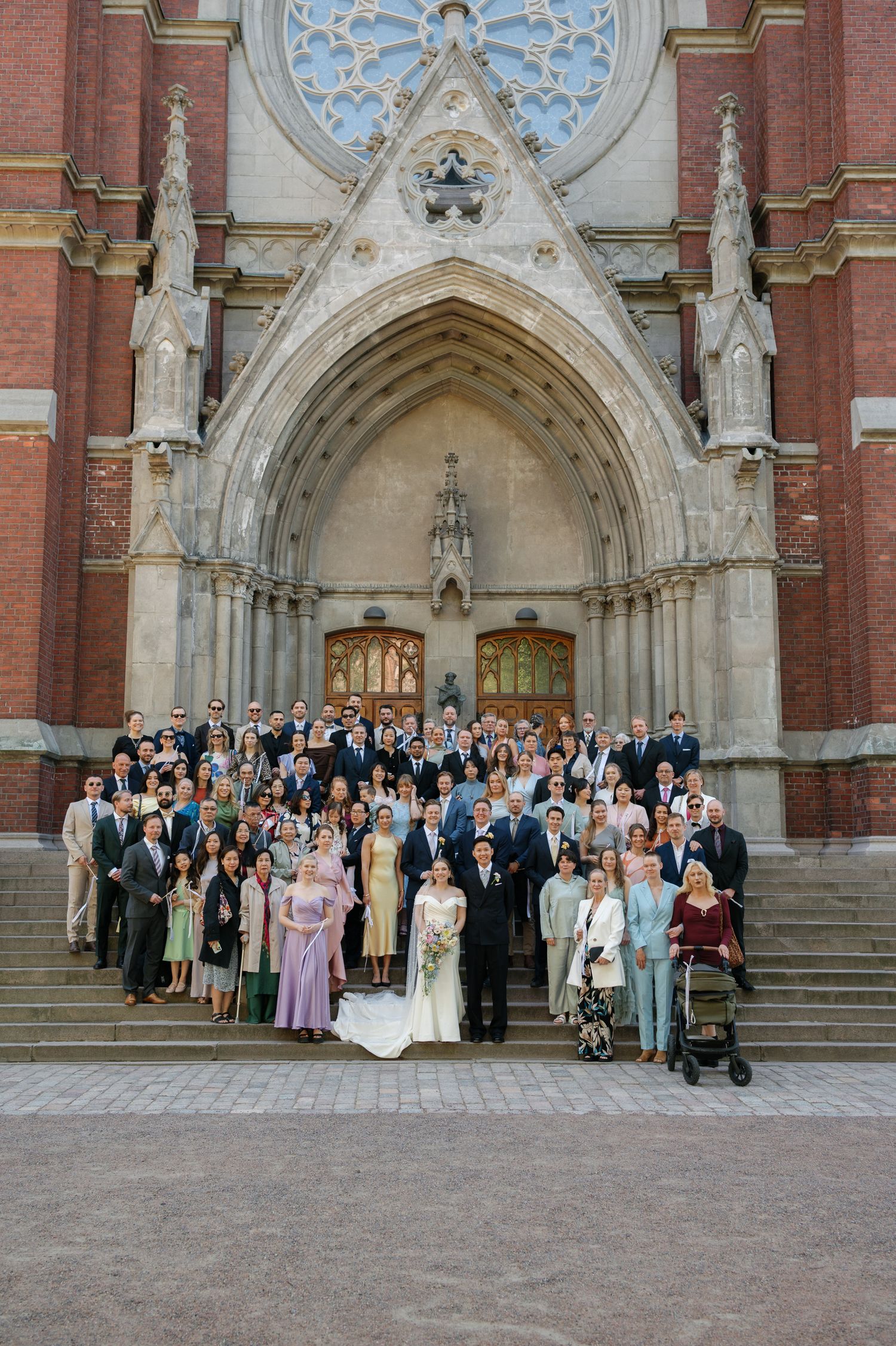 Large wedding party posing on steps of ornate Gothic brick church with arched doorway and rose window.