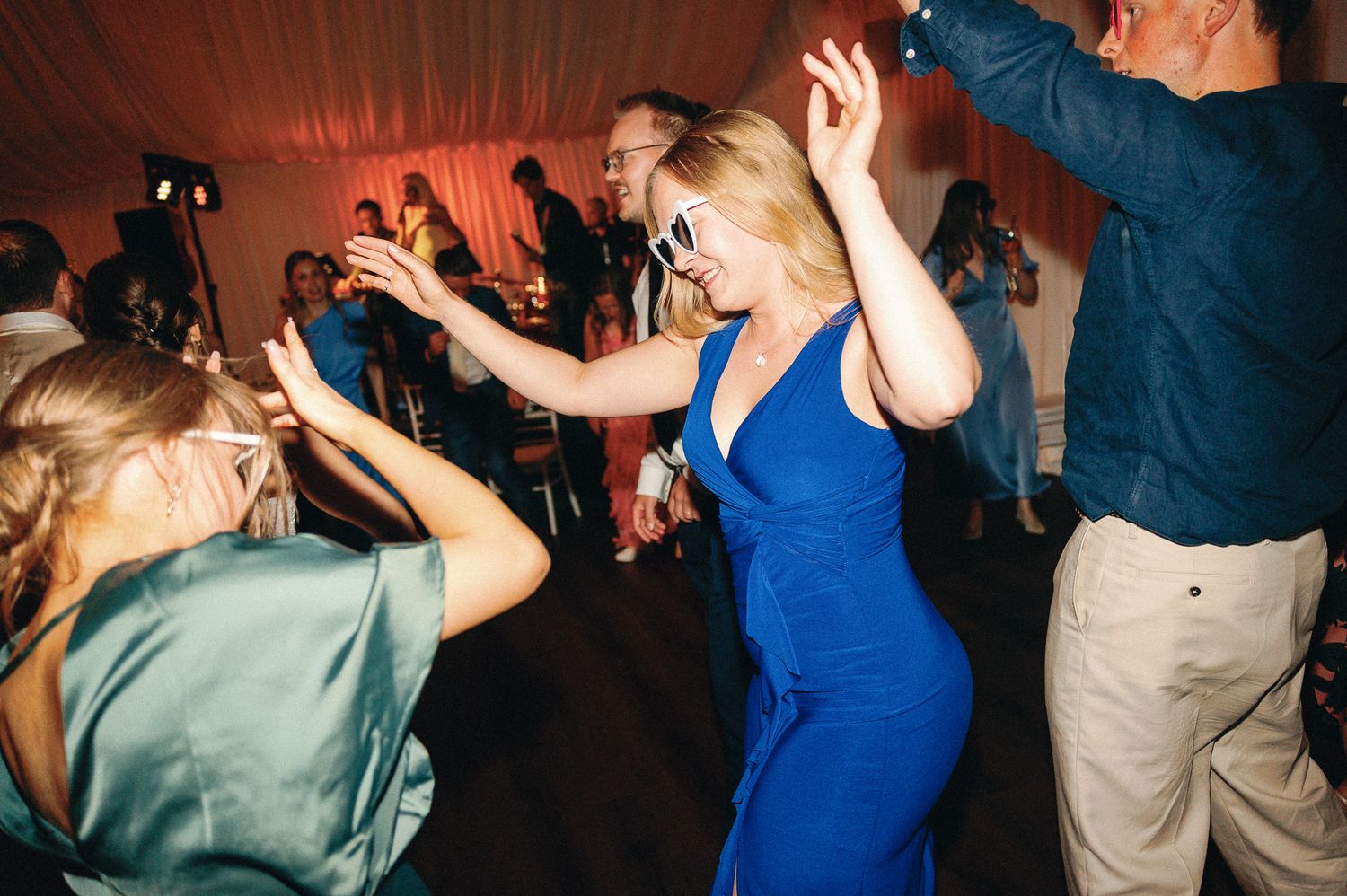 Woman in blue dress and white sunglasses dancing joyfully at a wedding reception party.