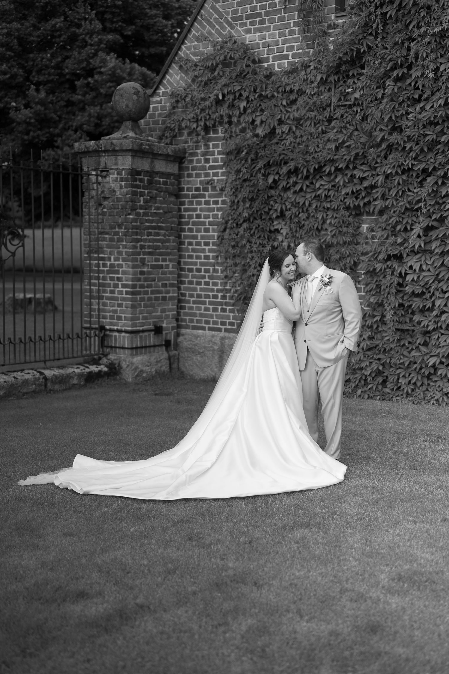 Black and white wedding photo of bride and groom sharing a romantic moment by an ivy-covered brick wall.