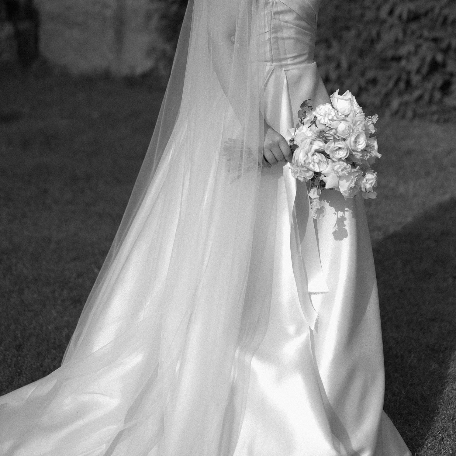 Black and white photo of a bride in a flowing satin wedding gown holding a bouquet of roses outdoors.