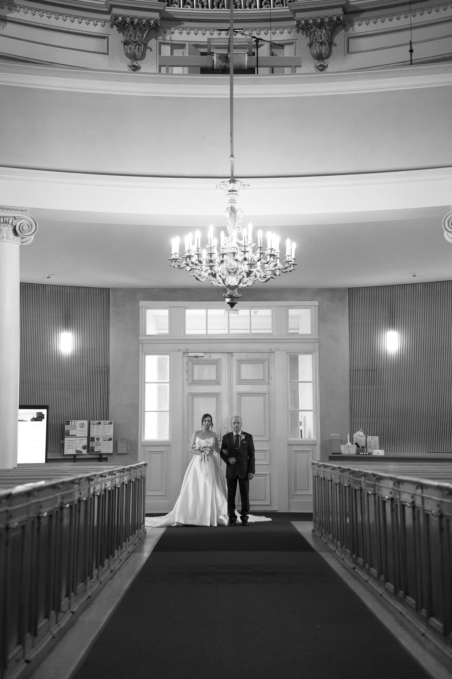 Black and white wedding photo of bride and groom standing at church entrance beneath elegant chandelier.