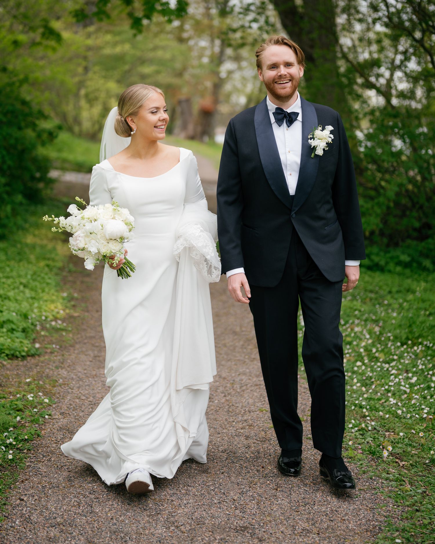 A bride in a white gown and groom in a black tuxedo walk hand in hand along a tree-lined path on their wedding day.