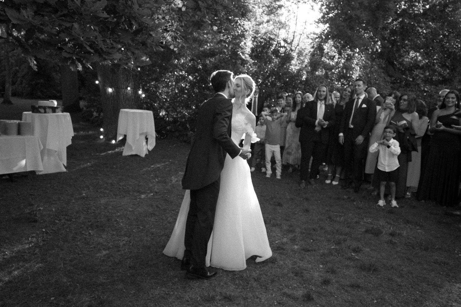 Black and white photo of bride and groom sharing first dance outdoors at evening wedding reception with guests watching.