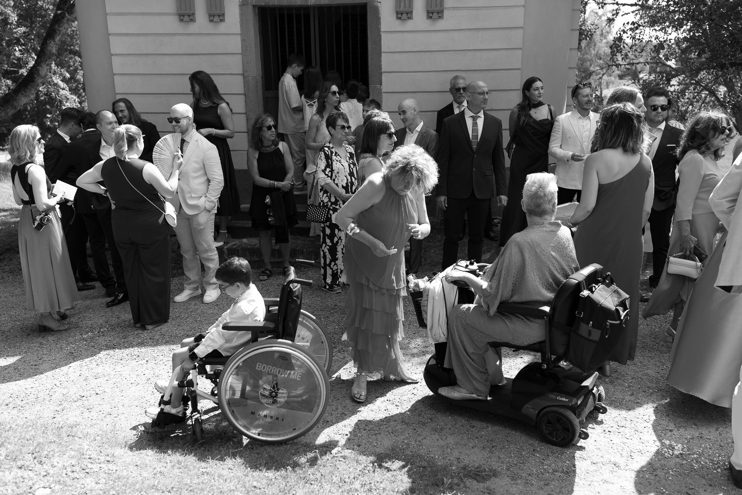 Black and white photo of wedding guests gathered outside a chapel, including elderly people in wheelchairs enjoying the celebration.