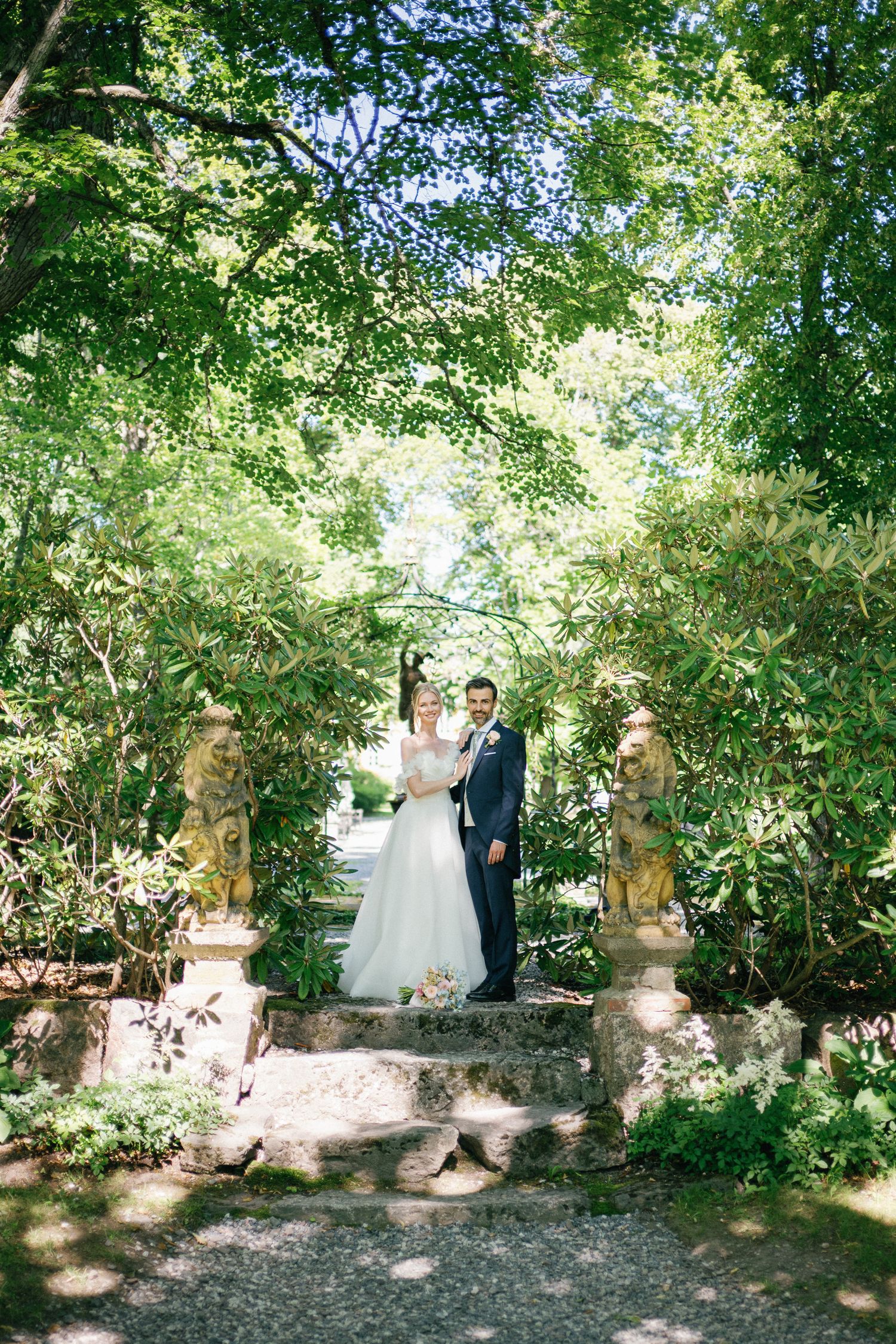 Bride and groom stand on stone steps surrounded by lush greenery and classical statues in a romantic garden setting.