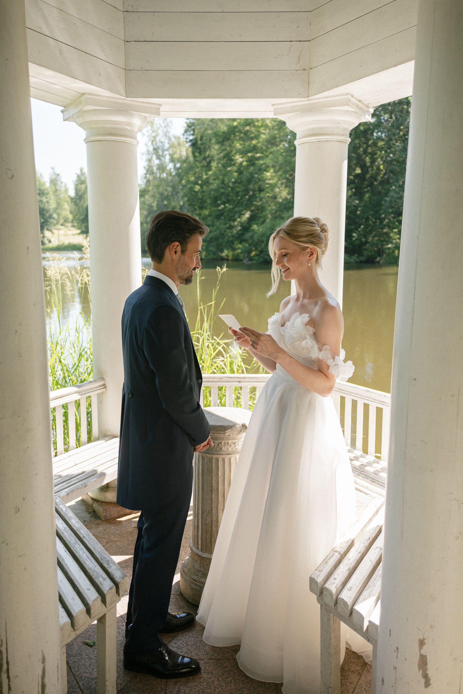 Bride reads wedding vows to groom in elegant white gazebo with columns, overlooking serene lake surrounded by lush greenery.